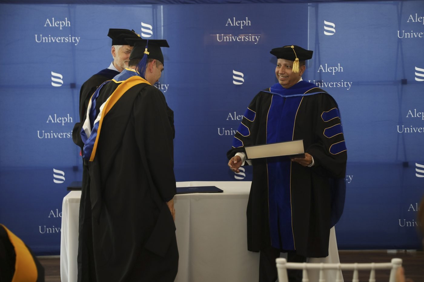 A graduate crossing the stage to receive their diploma in front of the Aleph University backdrop.